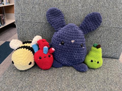 a yellow and black crocheted bee next to a crocheted red anatomical heart, next to a navy blue crocheted bunny, and a crocheted green pear