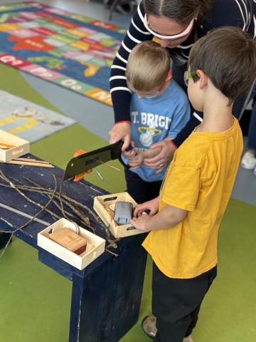 An adult helps a preschool-age child use a handsaw while another child watches.