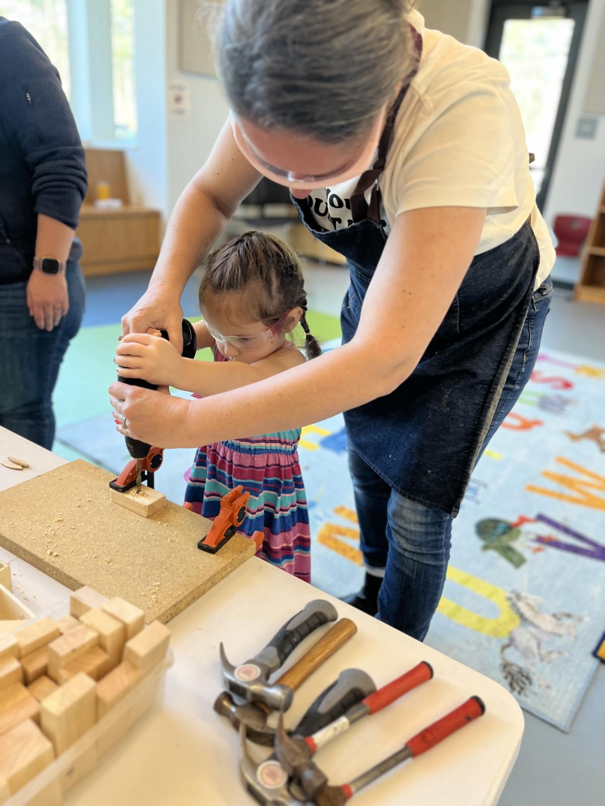 The teacher helps a preschool age girl use a power drill on a piece of wood. 